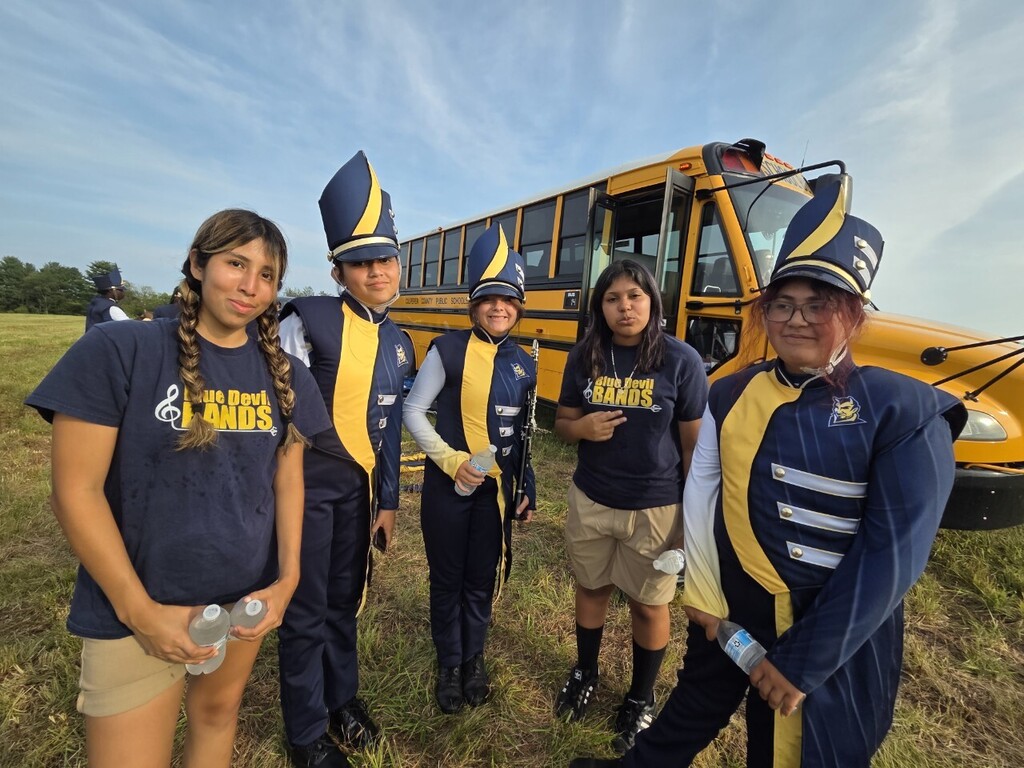 Five individuals in matching uniforms pose in front of a yellow school bus in a grassy field.