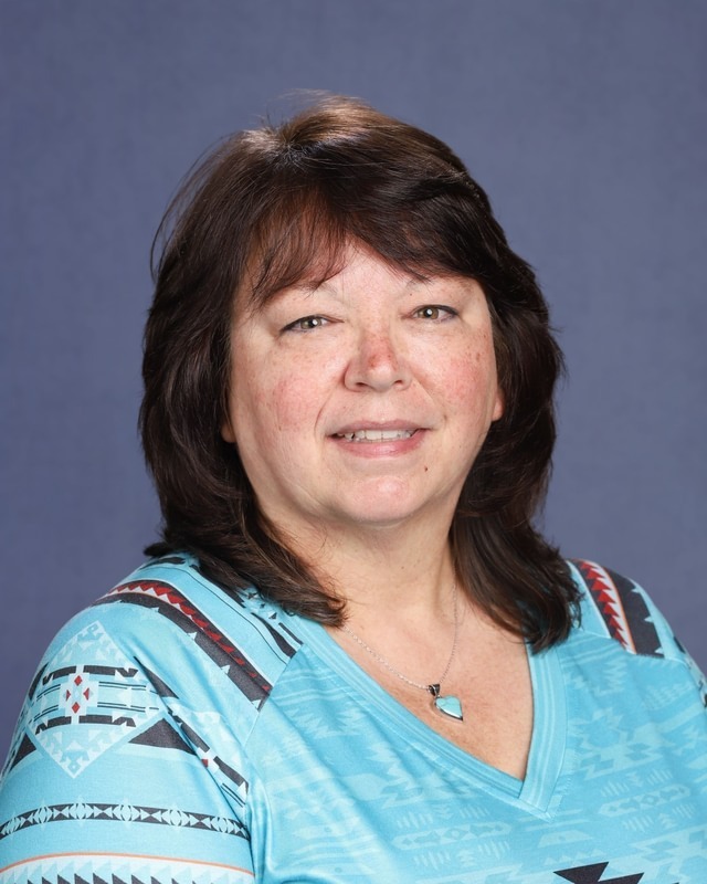 A woman with short brown hair smiles at the camera. She wears a blue top with a geometric pattern and a blue pendant necklace.
