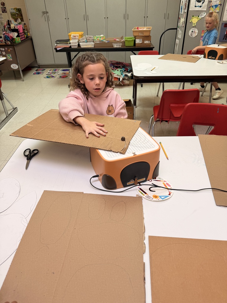 A student holds a large piece of cardboard over a cutting machine while working on a classroom art project, with additional cardboard pieces and art supplies spread across the table.