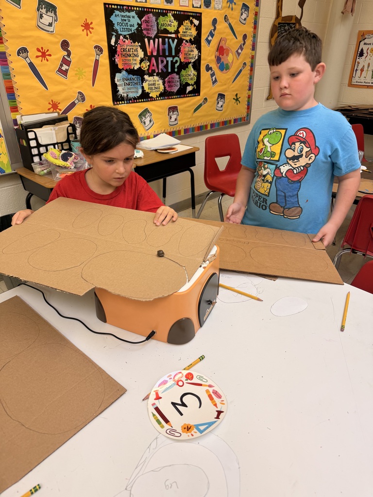 Two students work with large pieces of cardboard placed over a cutting machine during an art classroom project.