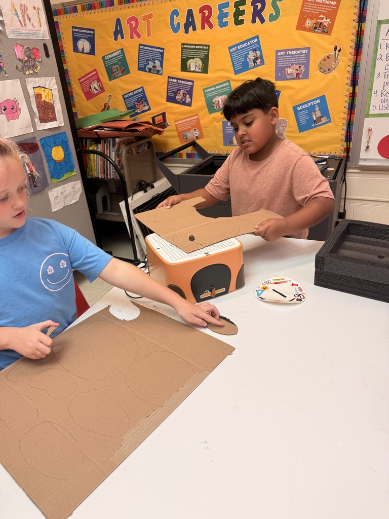 Two students work together with cardboard pieces around a cutting machine in an art classroom, with an “Art Careers” bulletin board in the background.