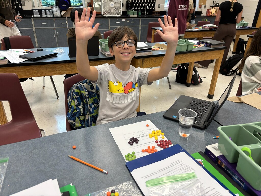 A boy wearing glasses raises both hands while sitting at a desk with colorful beads, papers, and a pencil.