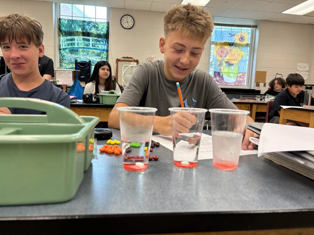 Two students in a classroom lab, one writes on paper while the other looks at cups of colored liquid.
