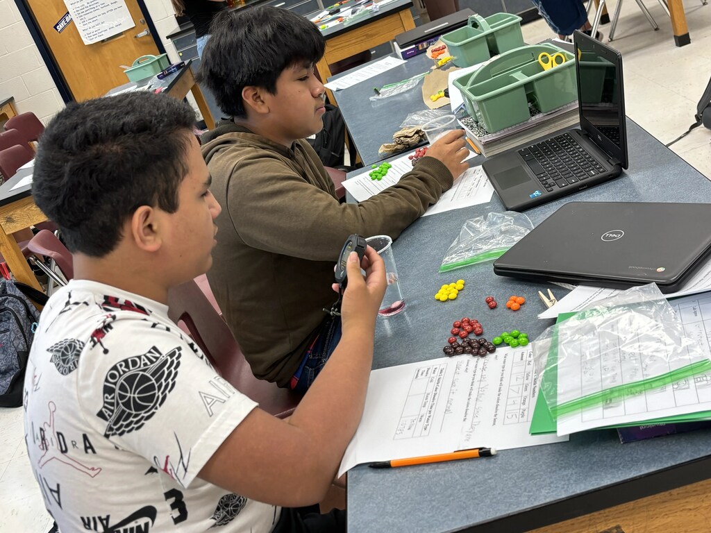 Two boys in a classroom are seated at a desk, using laptops and sorting colorful objects.