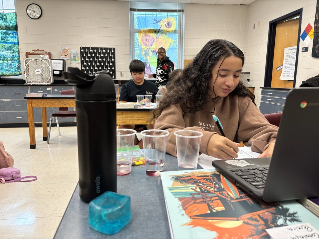 A person is sitting at a desk with a laptop, books, pens, and a water bottle.