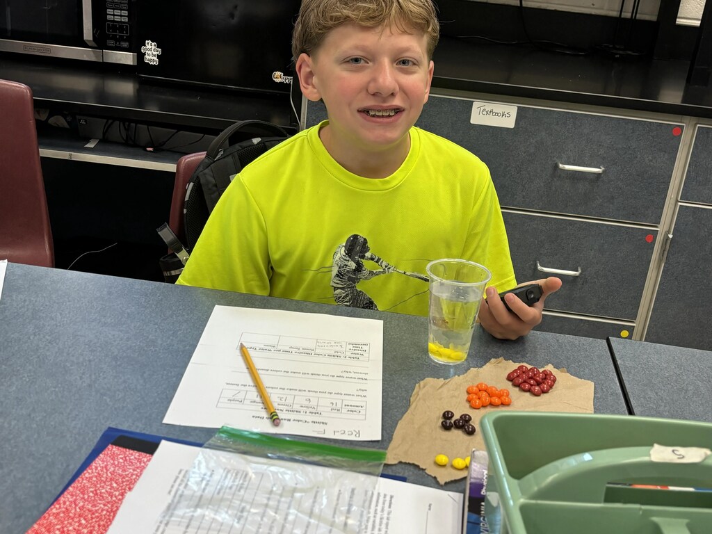 A young boy in a yellow shirt sits at a table with a glass of yellow liquid, a pencil, and a green container.