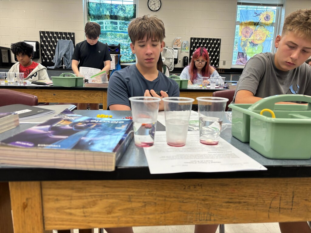 Students in a classroom, seated at tables with lab materials, looking down at a book and cups.
