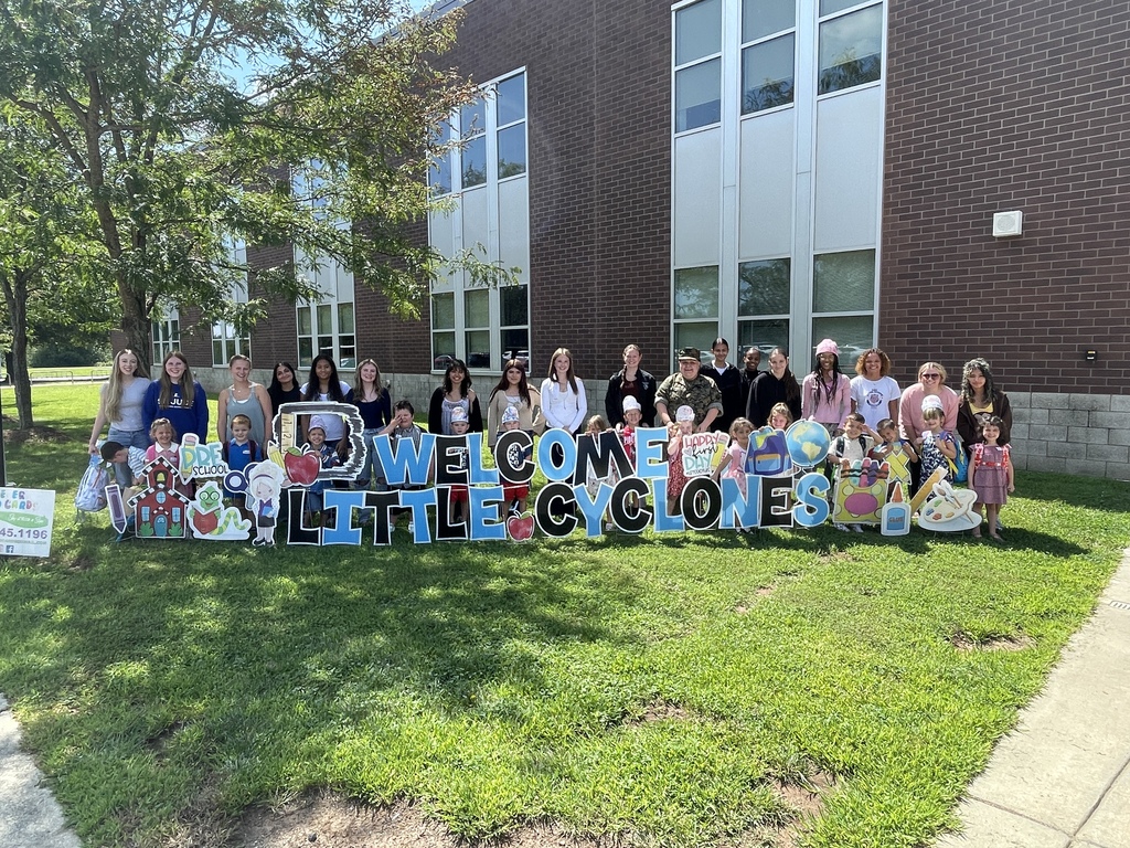 Group of people in front of a school building, posing for a photo with a sign reading Welcome Little Cyclones.