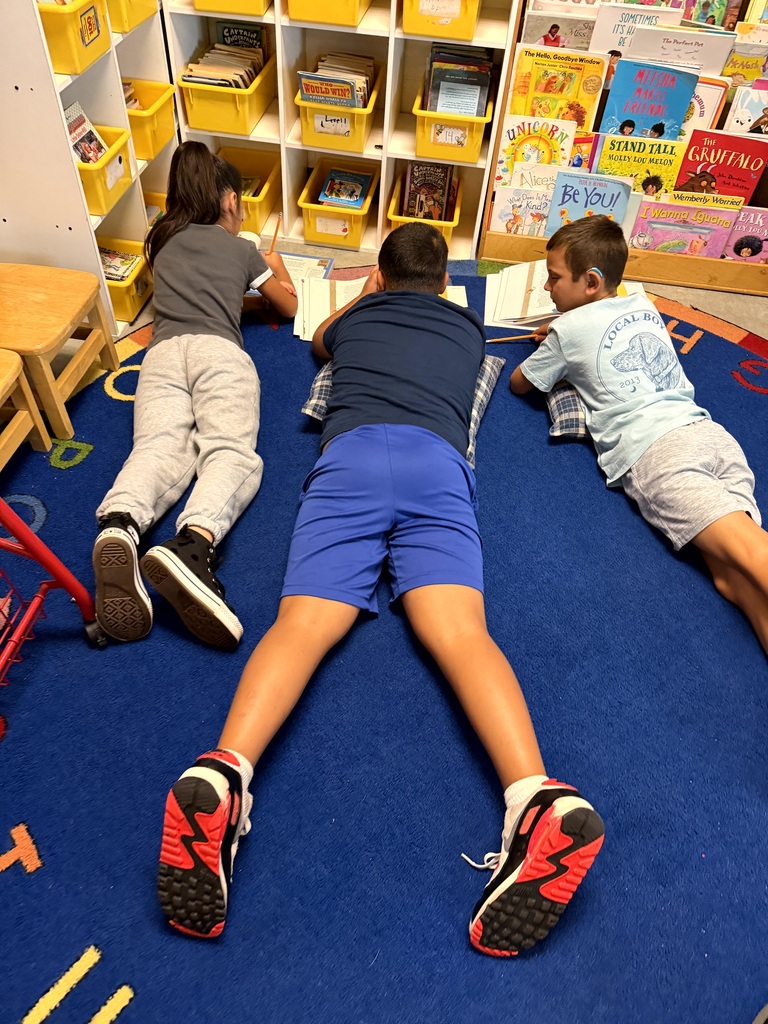 Three elementary students lie and sit on a classroom floor while reading and completing workbook activities together.