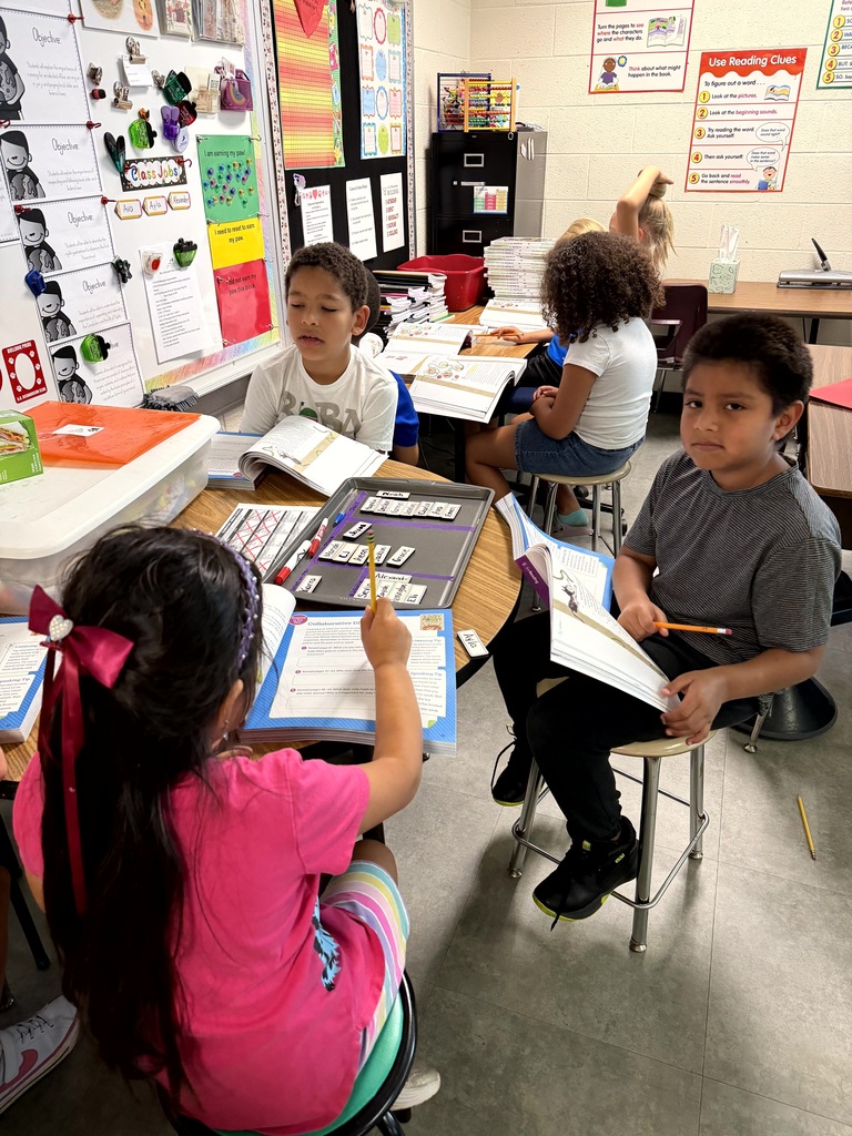 A small group of elementary students work with open books and pencils at tables during a classroom reading activity.