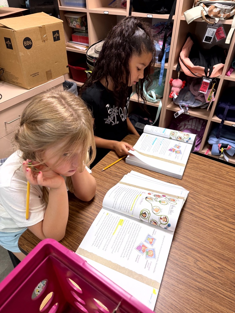 Two elementary students work at a classroom table with open reading workbooks, pencils, and highlighted passages.