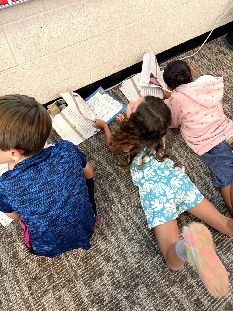 Three elementary students lie and sit on a classroom floor while reading and completing workbook activities together.