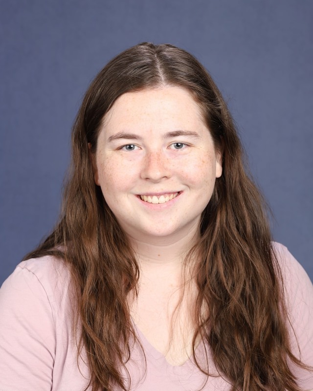 Woman with long brown hair, wearing a pink shirt, smiles and poses against a blue background.