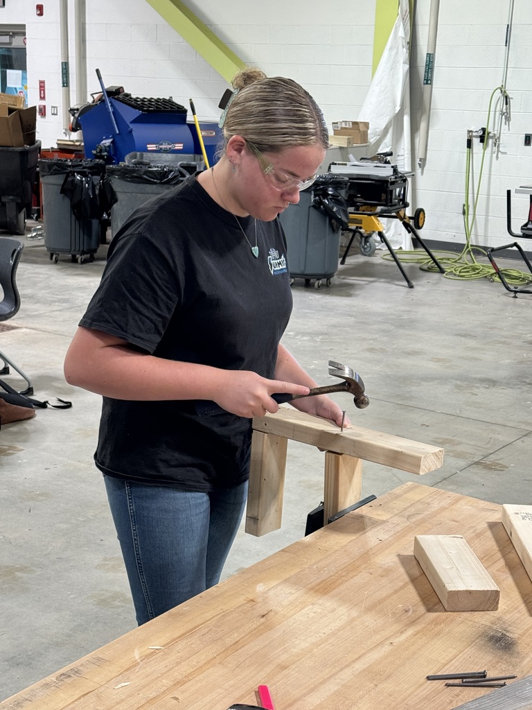A woman in a black t-shirt and glasses hammers wood on a workbench in a large room.