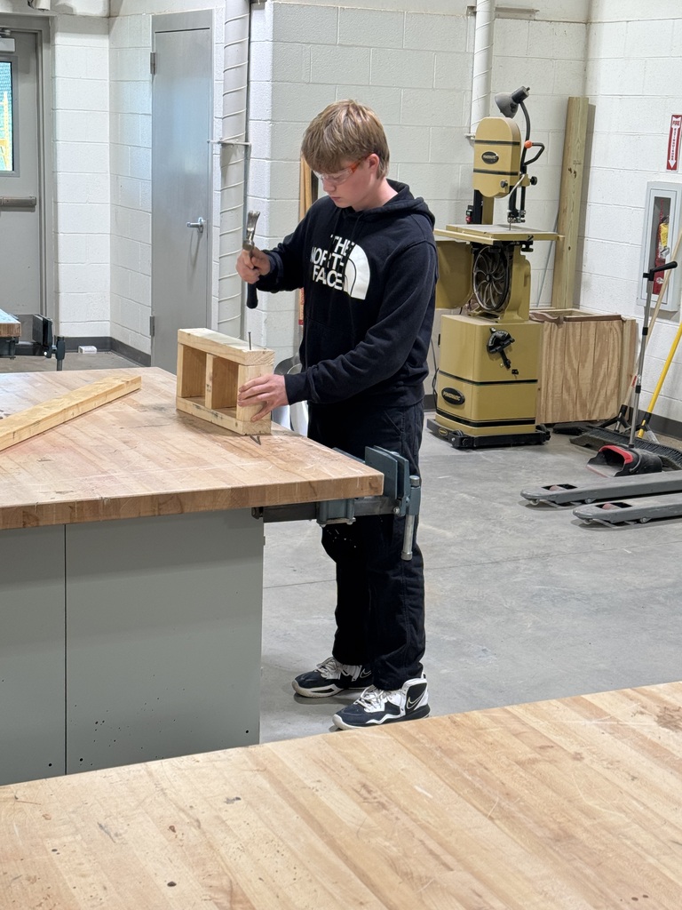 A person uses a hammer to put together a wooden box in a workshop. Behind him, there are workbenches and a yellow machine.