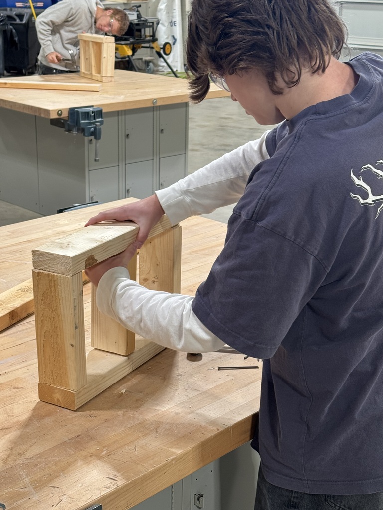 A person wearing a shirt assembles a wooden frame in a workshop. A wooden table is visible behind them.