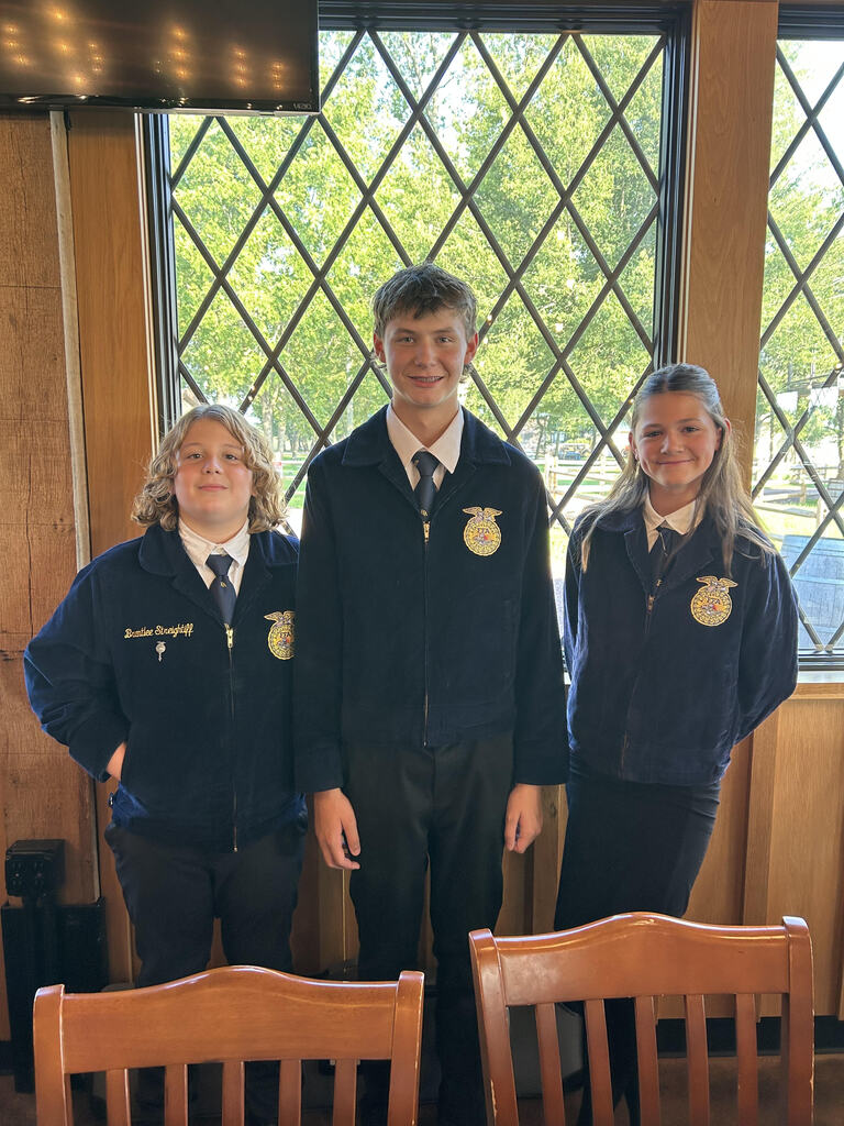 Three CMS FFA officers stand together indoors wearing official blue FFA jackets. From left to right are Secretary Brantlee Streightiff, Treasurer Camden Cox, and Historian Jocelyn Ezzell. The students are posed in front of large windows overlooking a sunny, tree-filled outdoor area.