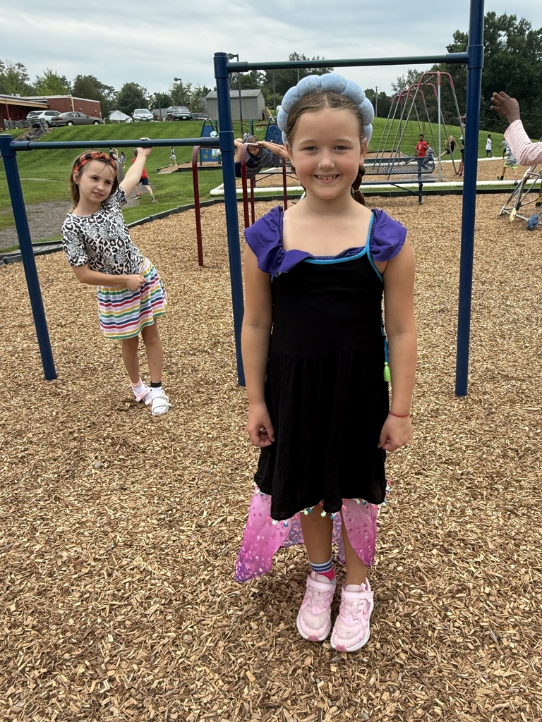 An elementary student wearing a colorful outfit and blue headband smiles on the playground, with another student in the background.