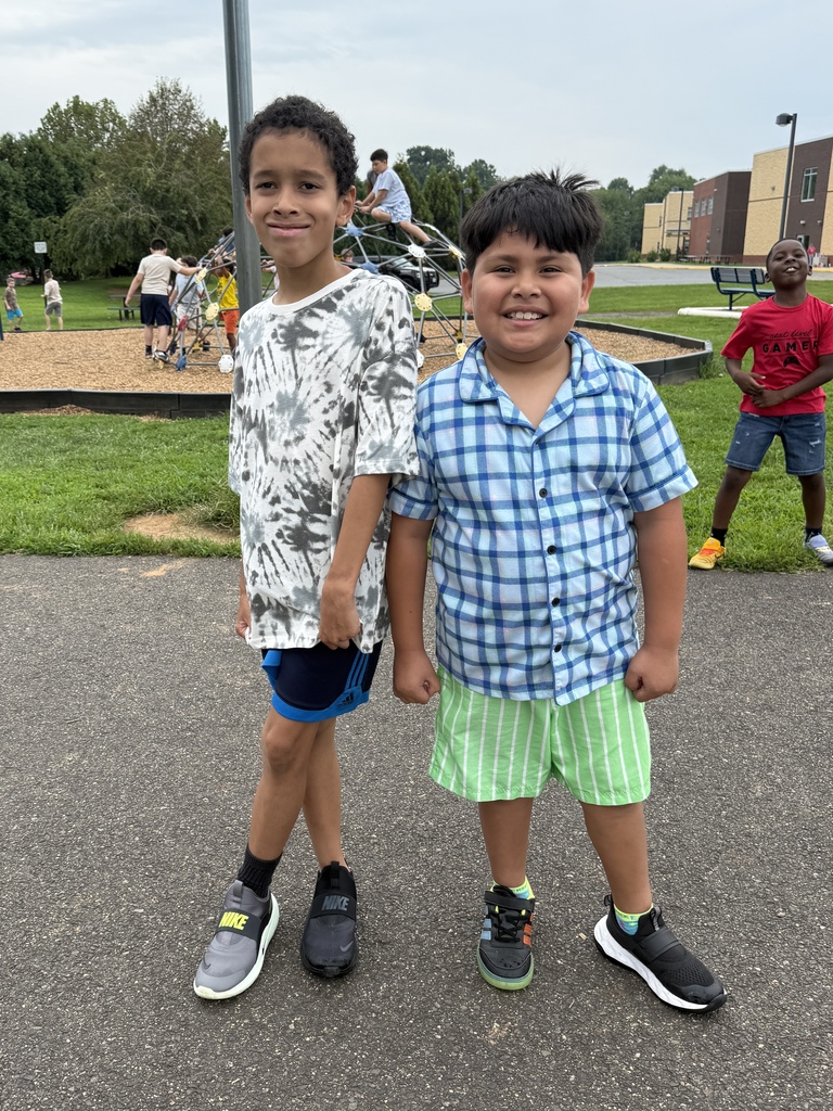 Two elementary students smile for a photo on the school playground while other students play in the background.