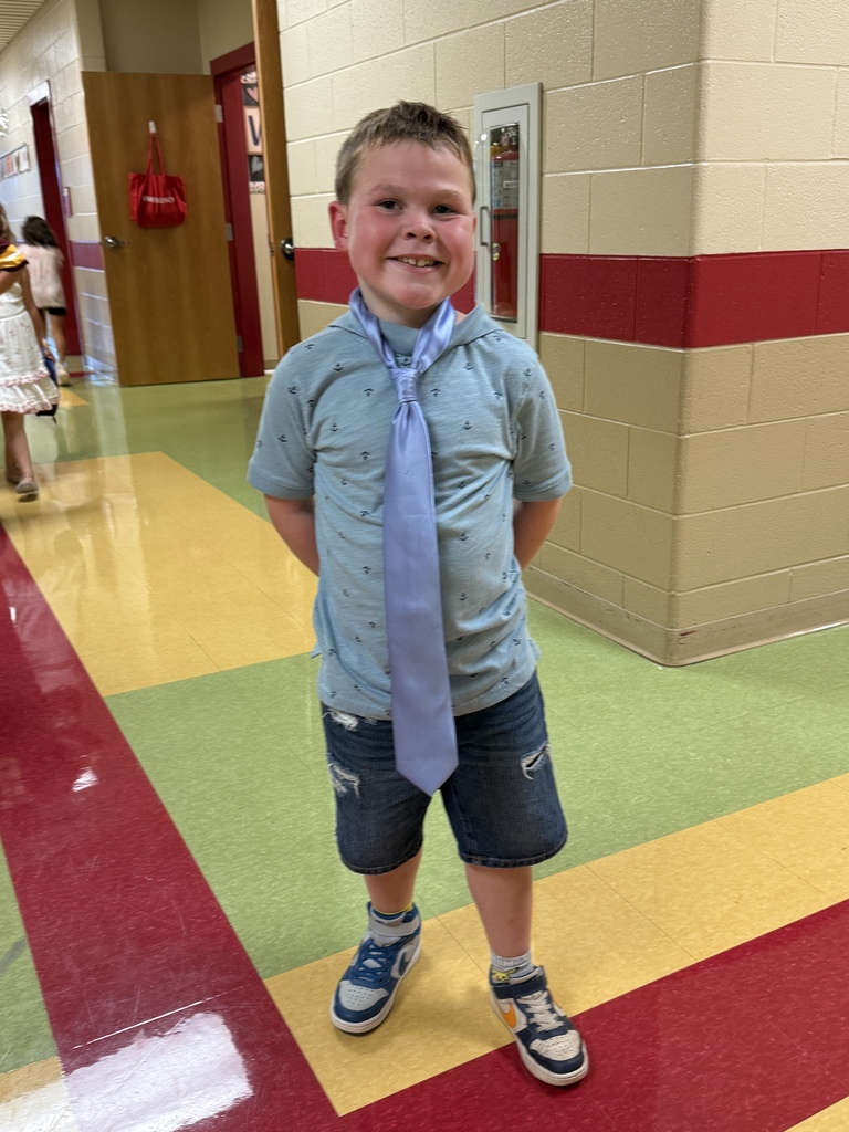 An elementary student wearing an oversized blue necktie smiles in a school hallway.