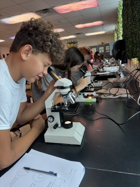 Several students examine objects with microscopes at a table. A pen rests on a white sheet of paper.