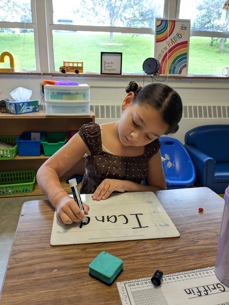 A girl sits at a desk writing the word "trace" on a white sheet. Behind her are shelves, windows, and chairs.
