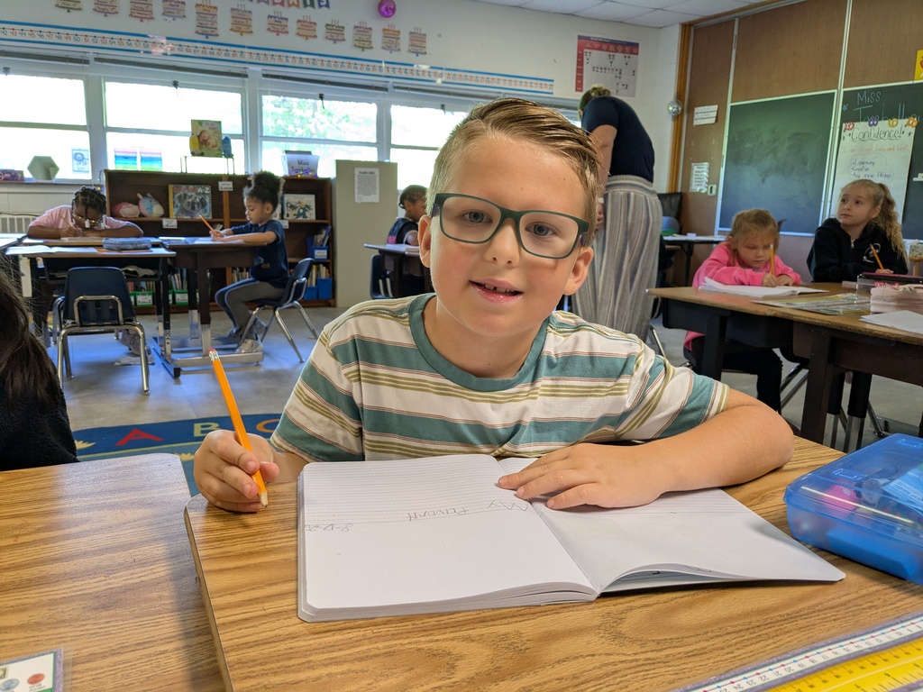 A boy in glasses sits at a desk, holding a pencil and writing in a notebook.