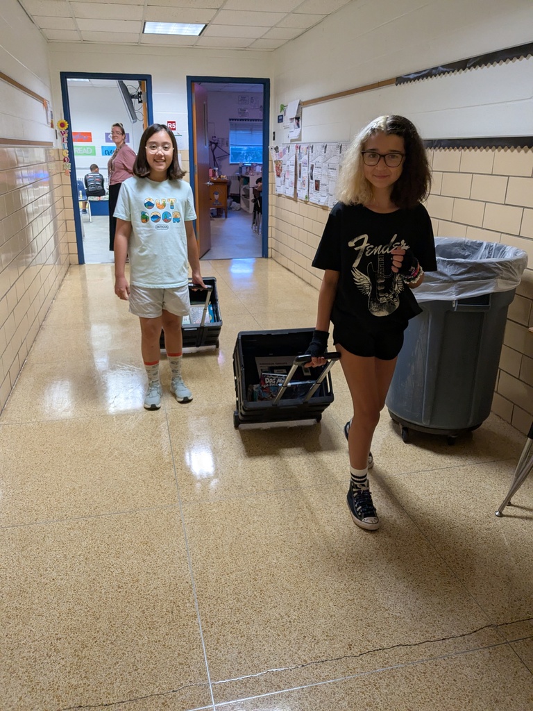 Two girls in a school hallway carrying wheeled boxes. One has glasses, the other wears a white t-shirt. A trash can and open door are in the background.
