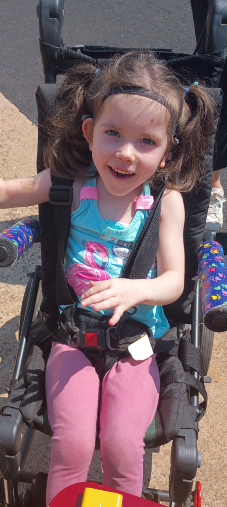 A young girl sits in a wheelchair, smiling, with a blue shirt, pink pants, and a black seat belt.