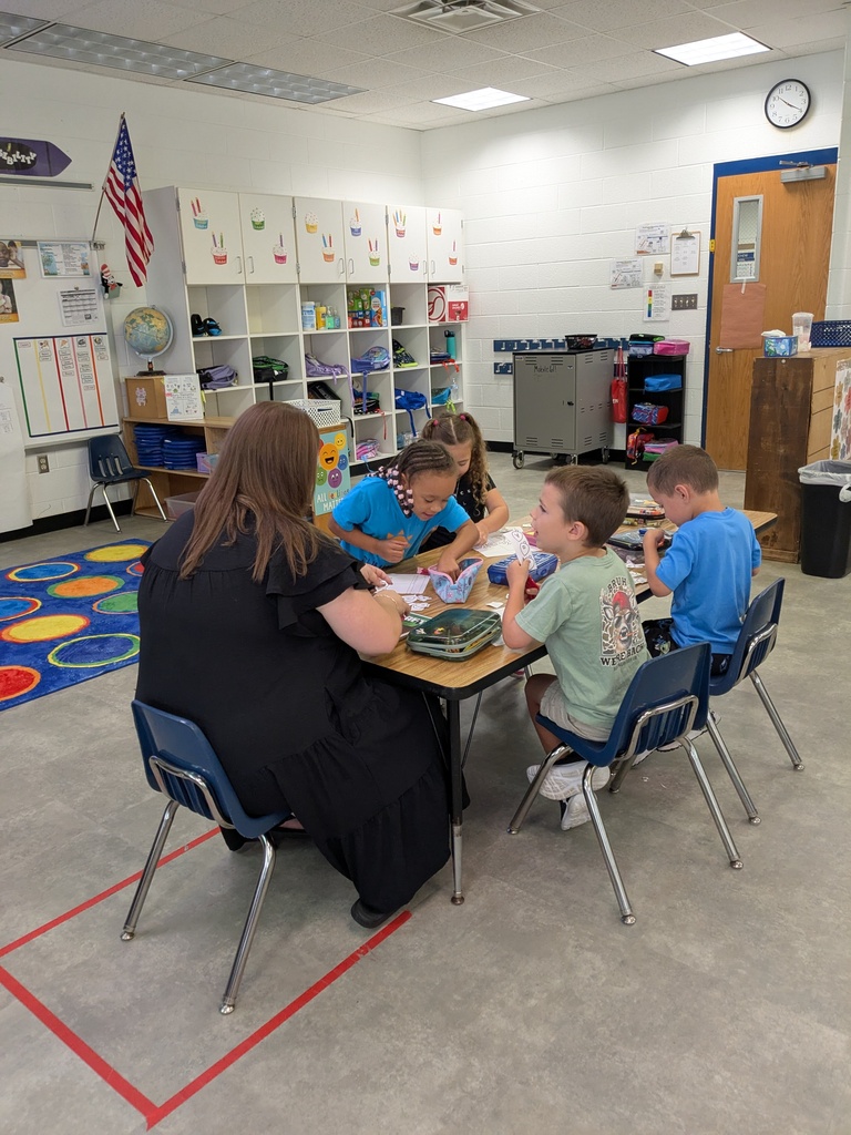 Teacher seated with four children at a table. Behind her, shelves, a globe, and an American flag.