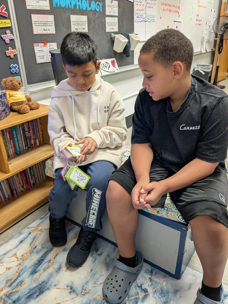 Two kids sitting on a bench, one holding cards. Behind them, a bookshelf and a whiteboard with various papers.
