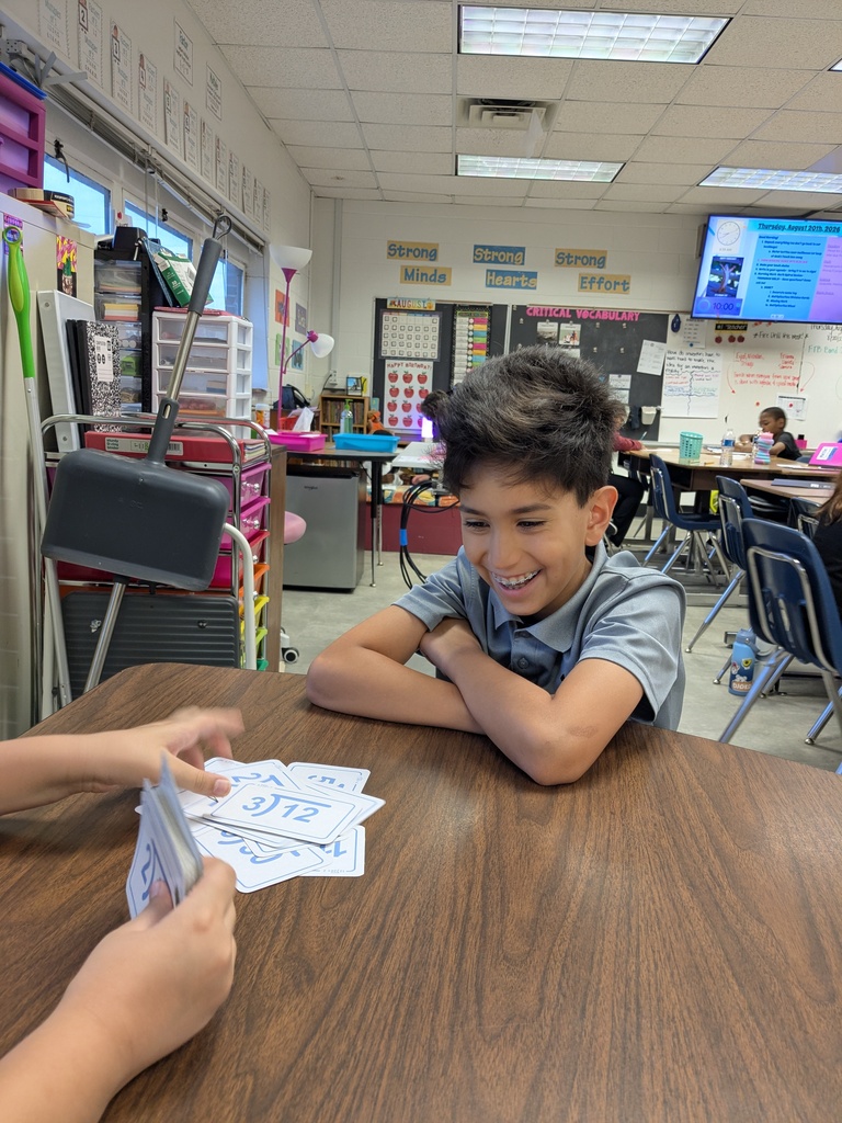 A boy sits at a table in a classroom with cards, smiling. Another person hands him a card.