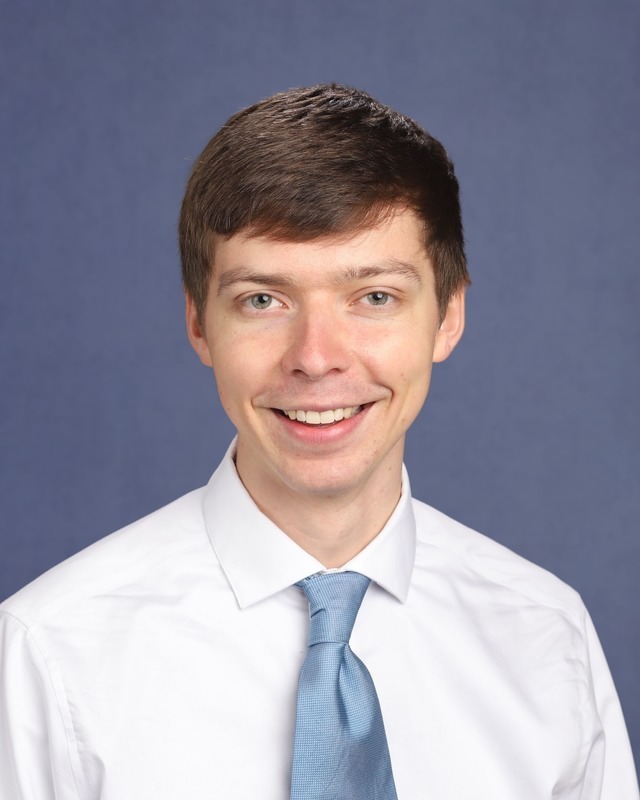 A person with short hair, wearing a white shirt and blue tie, smiles against a blue background.