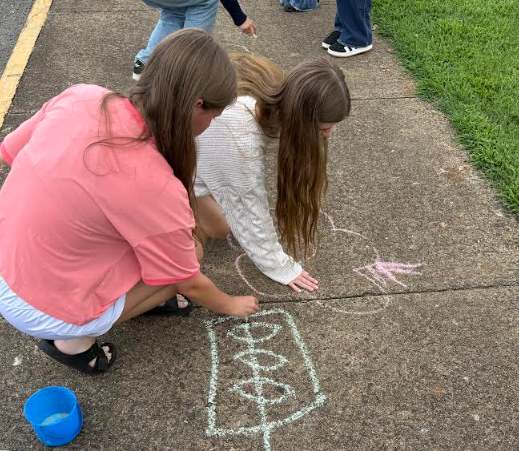 Two girls draw on a sidewalk with chalk. One wears a pink shirt and the other a white long-sleeve shirt.
