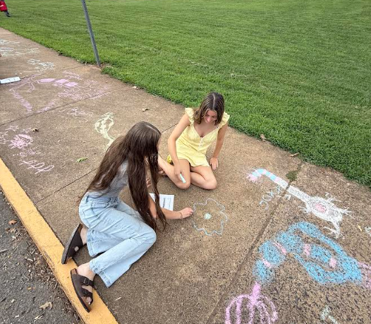 Two people on a sidewalk with chalk drawings. One person is kneeling and holding a piece of paper.