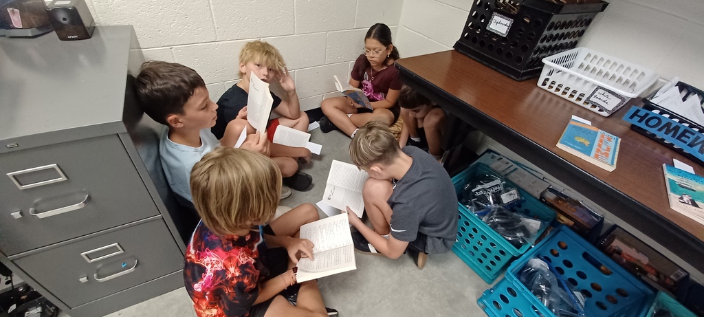 Six students sit closely together on the classroom floor near a desk and filing cabinet, reading books during a small-group activity.