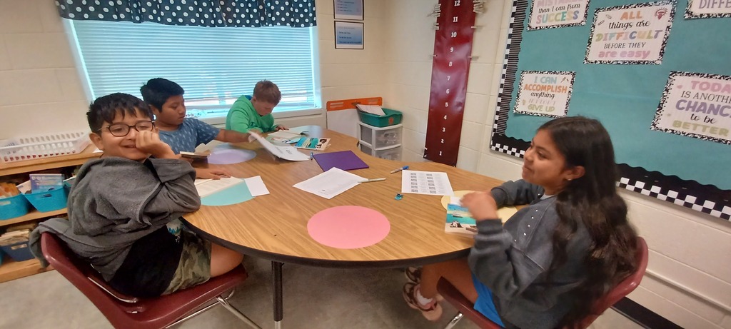 Four students sit around a classroom table with books, papers, and colorful materials while participating in a reading and learning activity.