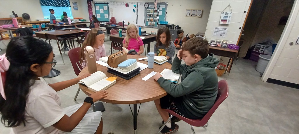 Five students sit around a classroom table reading books together, while other students work in small groups around the room.