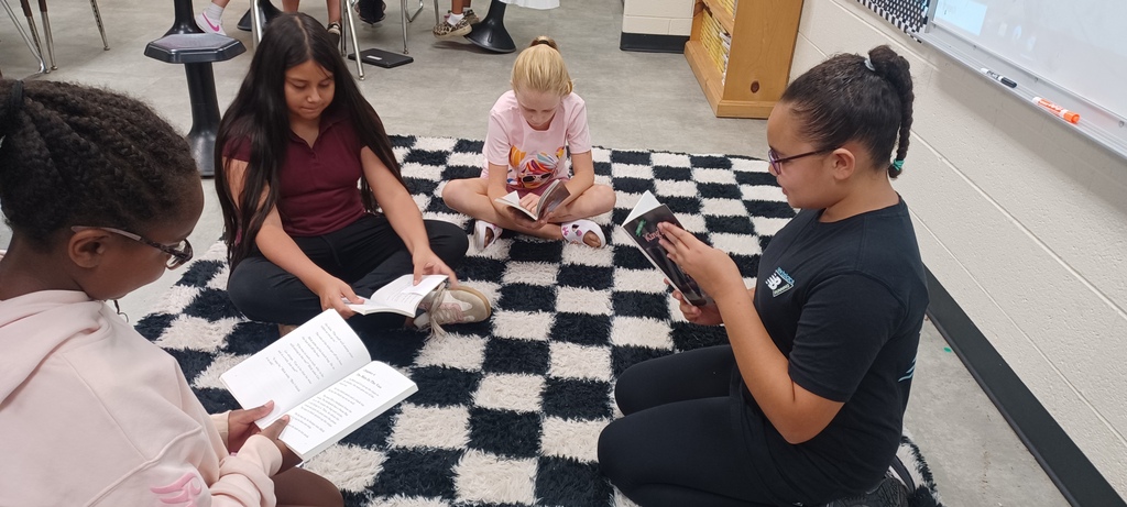 Four students sit together on a black-and-white checkered classroom rug, each reading a book during a small-group reading activity.