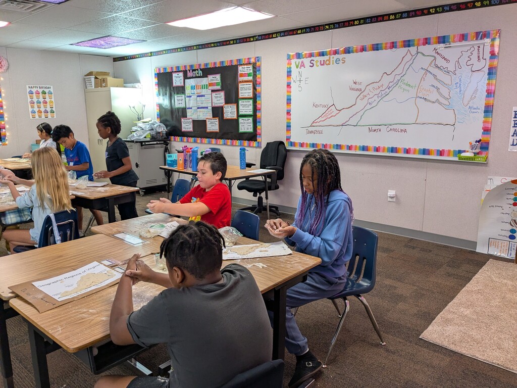 Classroom with several children seated at desks, engaged in activities. Whiteboard with drawings and charts in the background.