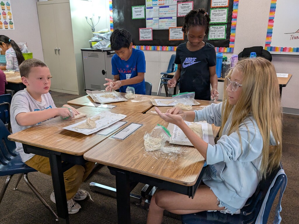 Kids are sitting at desks, working on a craft project. A girl wearing glasses holds up her work.