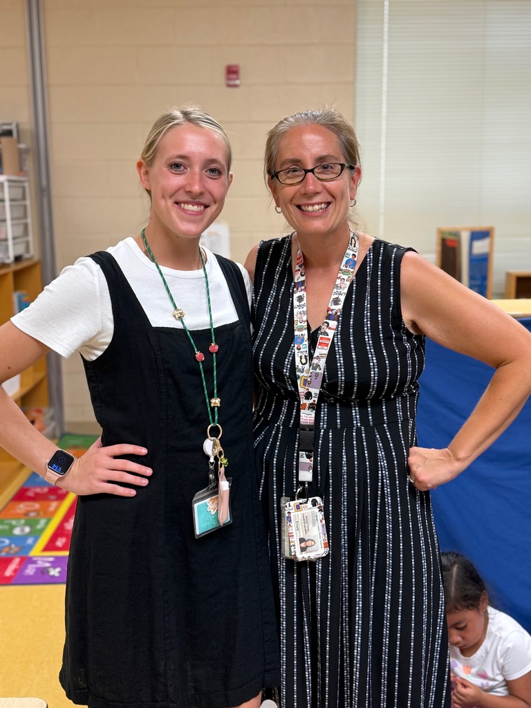 Two women stand together, smiling, in a classroom. One wears a white top and a necklace. The other wears a black dress with a lanyard.