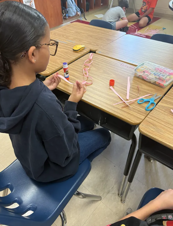 A student sits at a classroom table assembling a pink paper chain using paper strips, scissors, and glue. Photo 6: Two stud