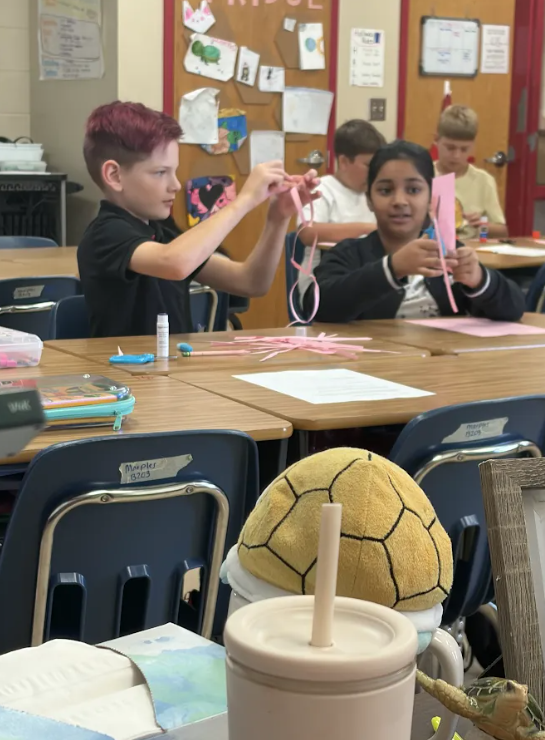 Students sit at classroom tables cutting and connecting strips of pink paper to create paper chains for a STEM challenge.
