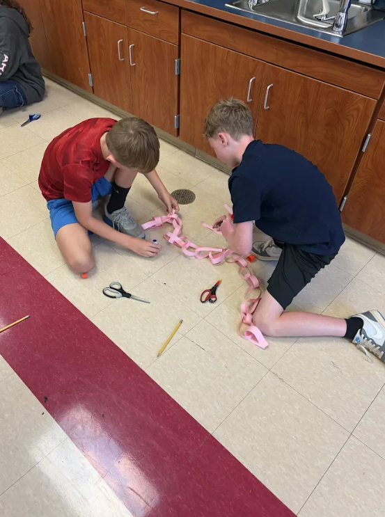 Two students work together on the classroom floor, using scissors and glue to construct a long pink paper chain.