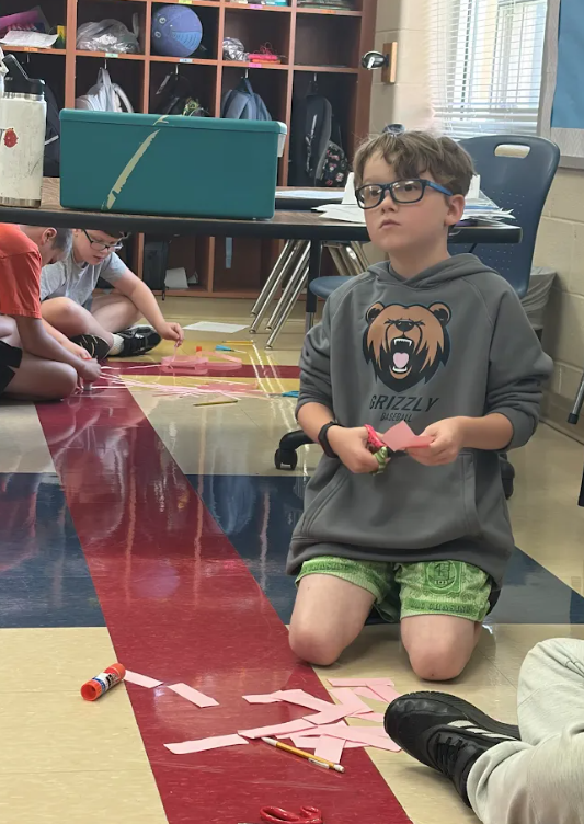 A student kneels on the classroom floor cutting strips of pink paper while classmates work on their paper chains in the background.
