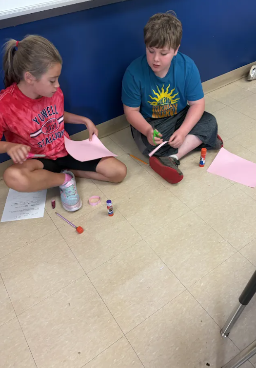 Two students sit on the classroom floor cutting pink paper into strips as they work together on a paper chain STEM challenge.