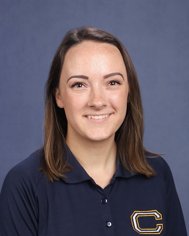 A woman with brown hair smiles at the camera. She wears a navy blue polo shirt with a gold "C" logo.