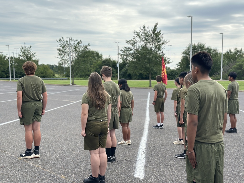 Group of people in green uniforms stand in a parking lot with trees and a cloudy sky.