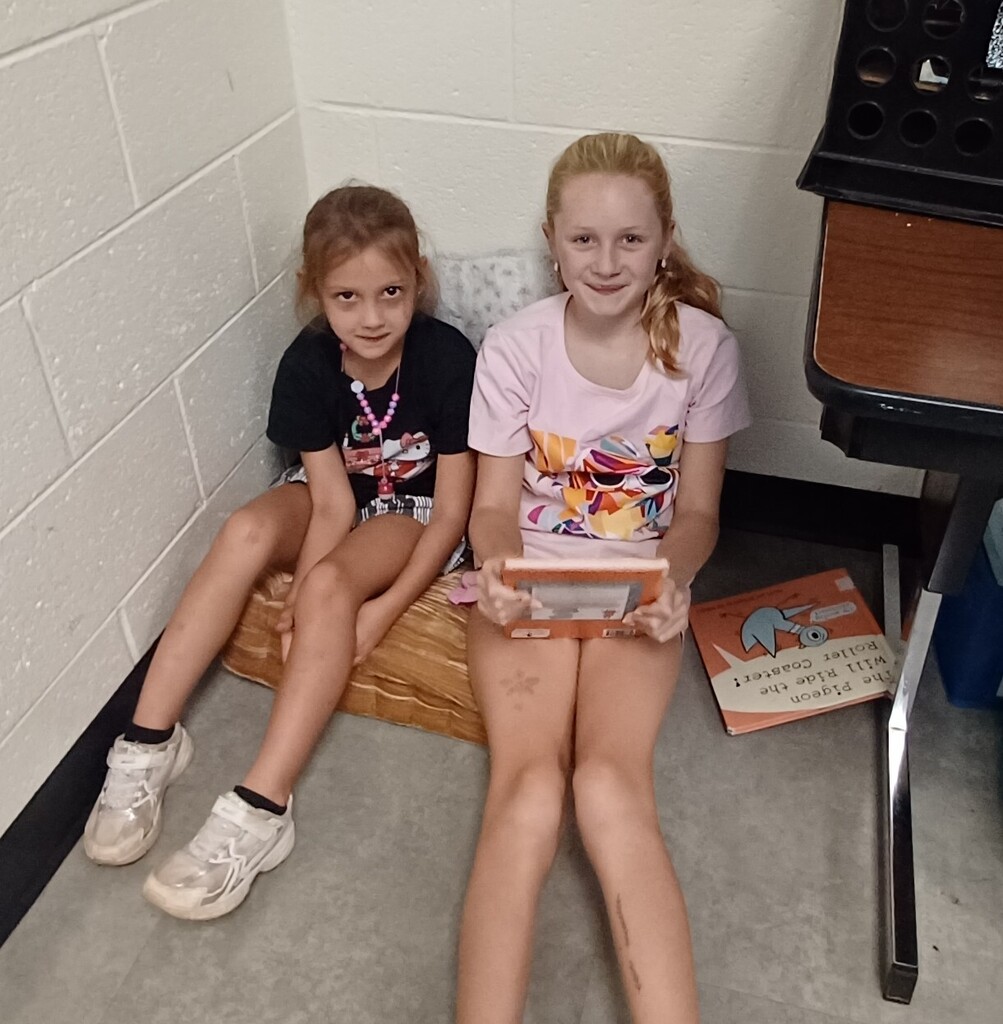 Two young girls sit on a wooden bench in a room with a white wall, holding a handheld game console.
